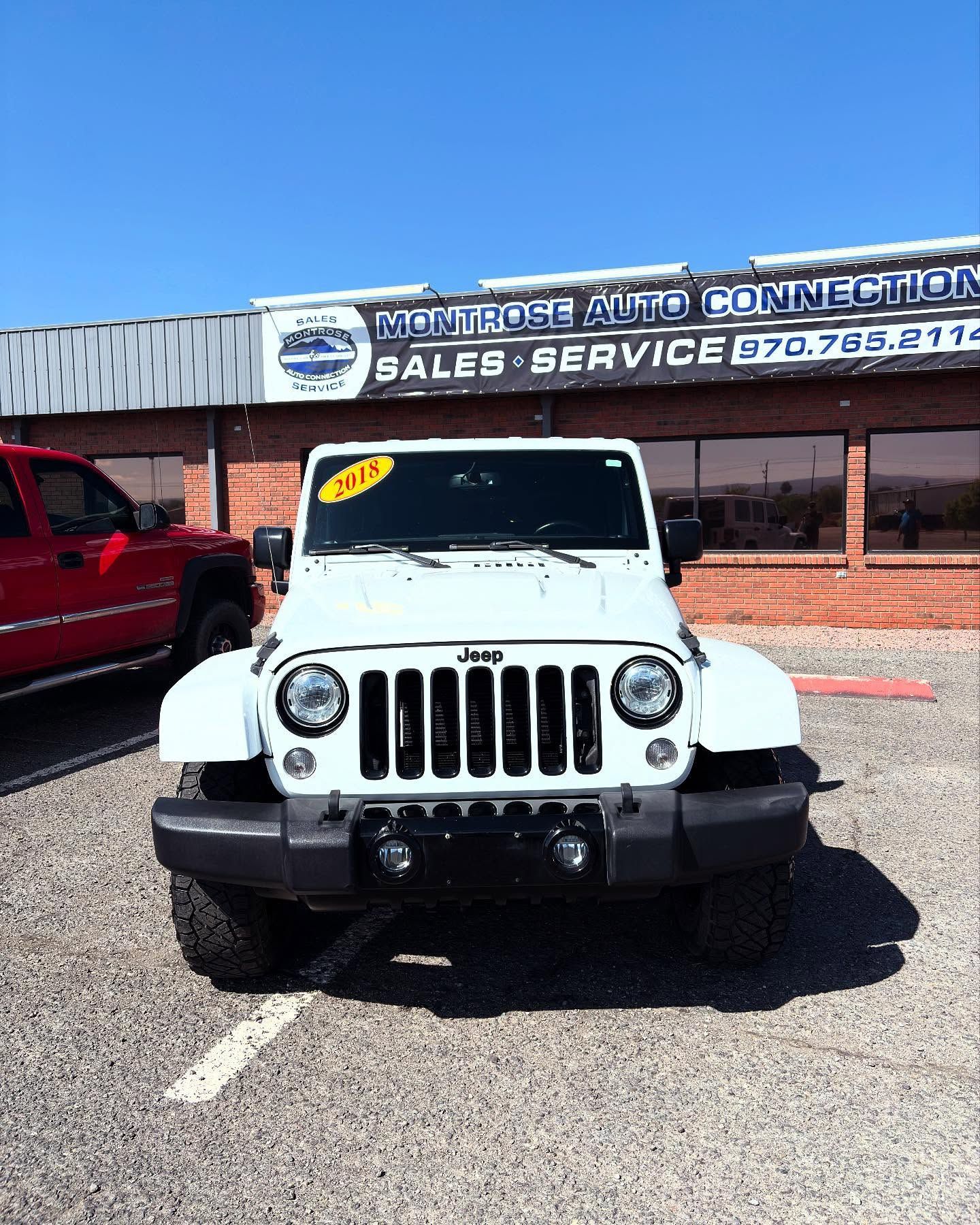 A white 2015 Jeep Wrangler parked in front of the Montrose Auto Connection dealership on a sunny day.