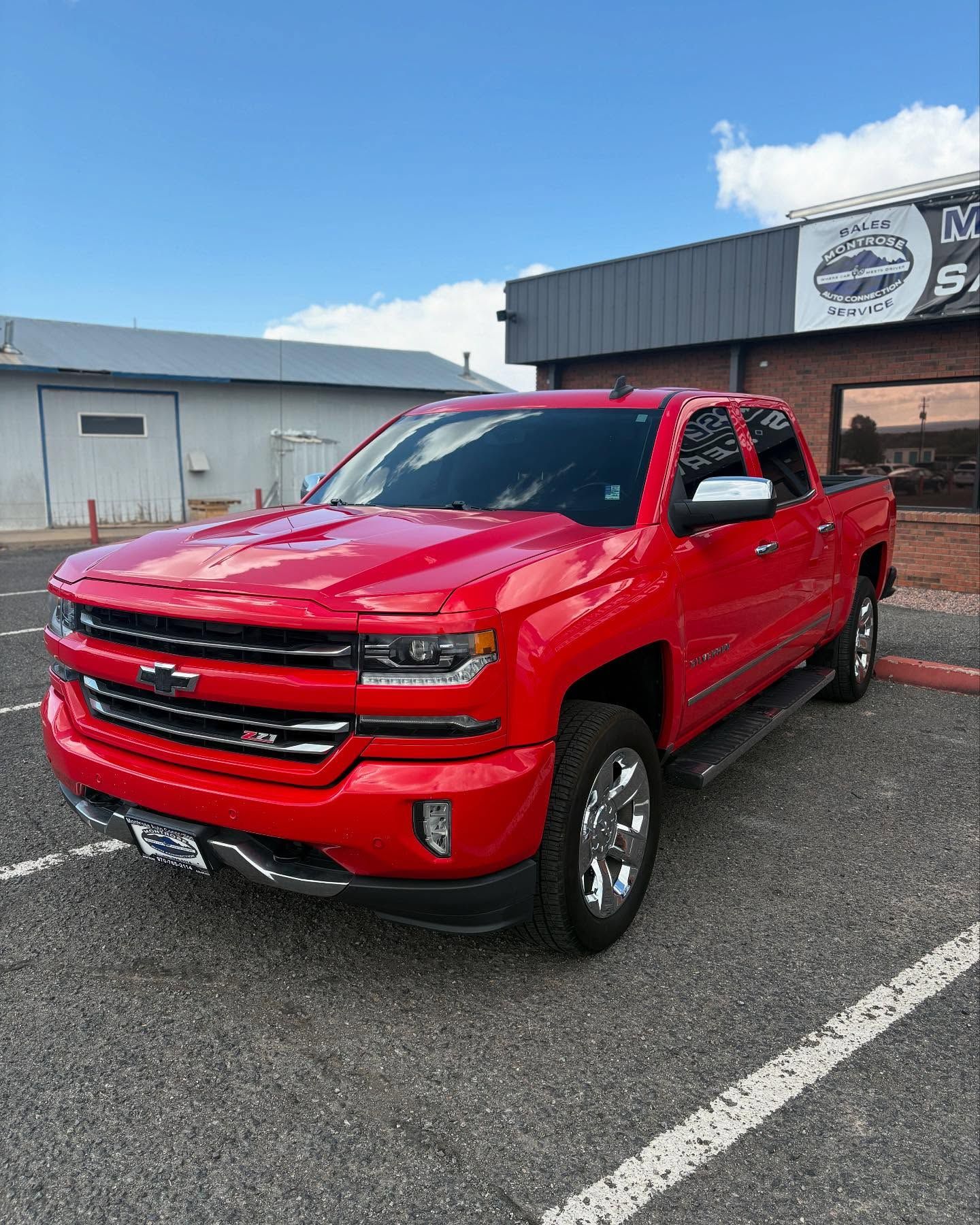 A bright red Chevrolet Silverado pickup truck parked in an outdoor lot in front of a building under a blue sky.