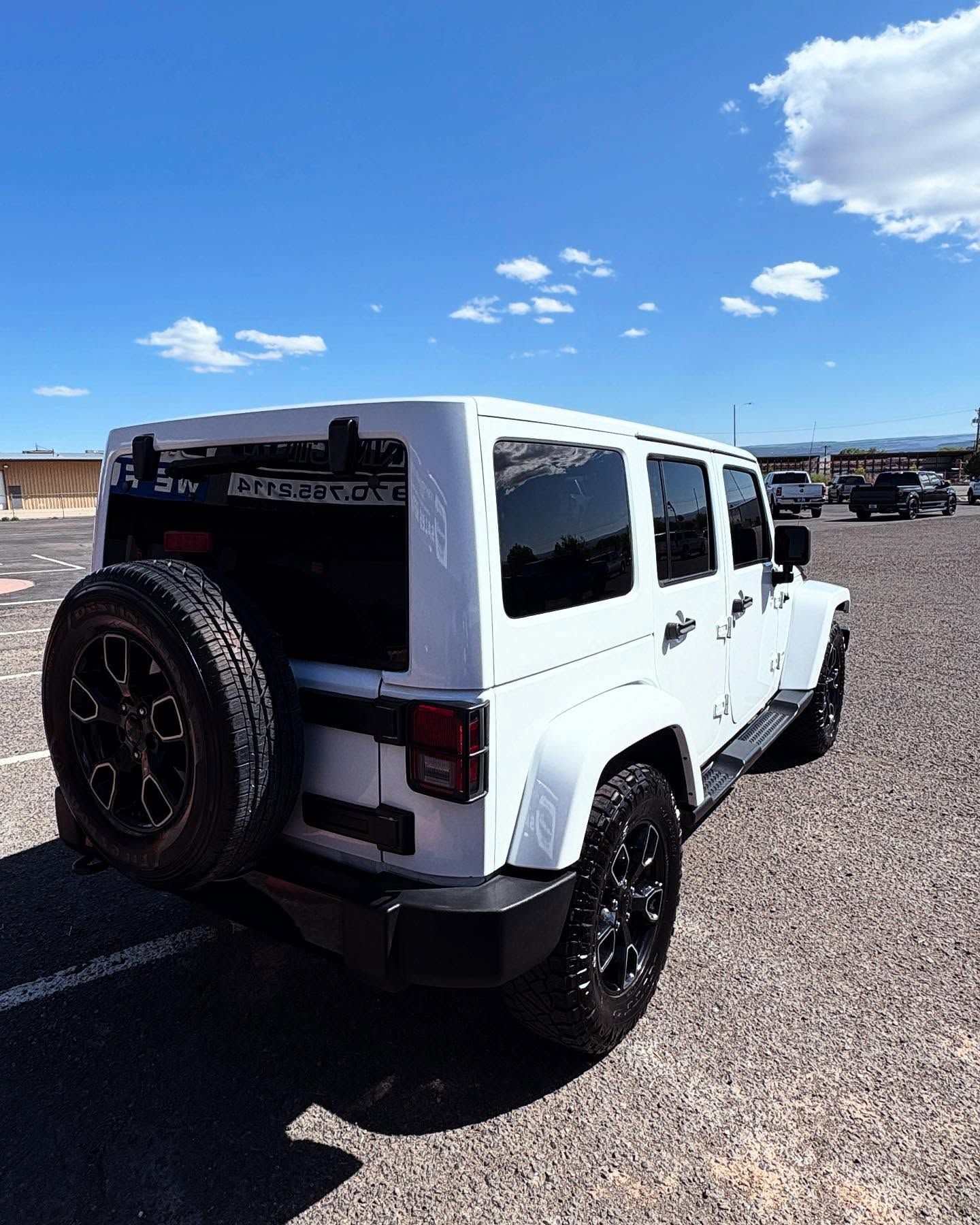 A white Jeep Wrangler parked on a gravel lot under a clear blue sky, viewed from the rear three-quarter angle.