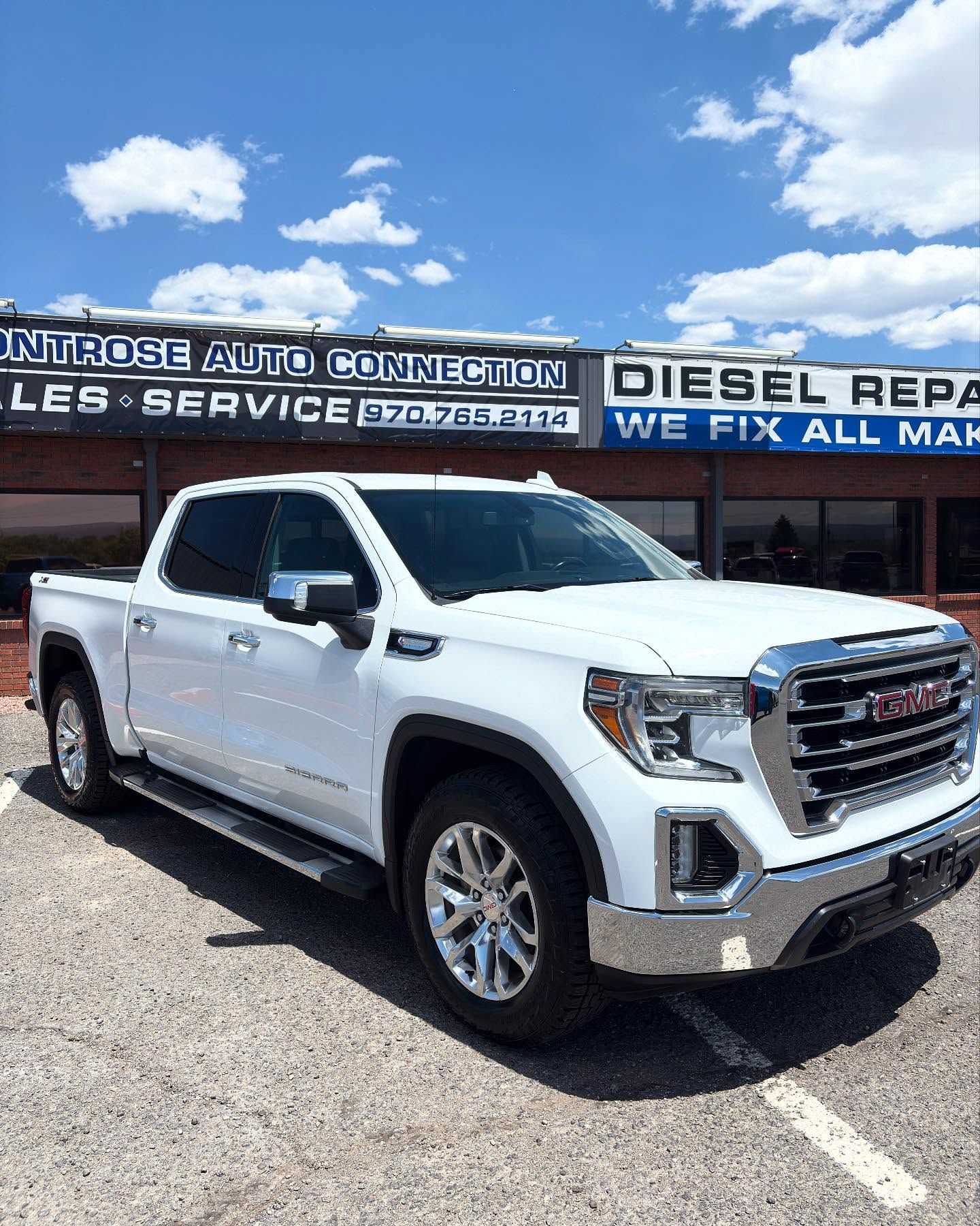 A white GMC Sierra pickup truck parked in front of the Montrose Auto Connection dealership under a blue cloudy sky.