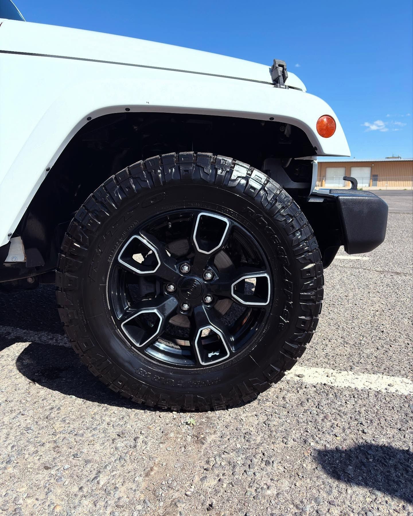 A close-up of the front driver-side wheel and fender of a white Jeep parked on a gravel lot under a clear blue sky.