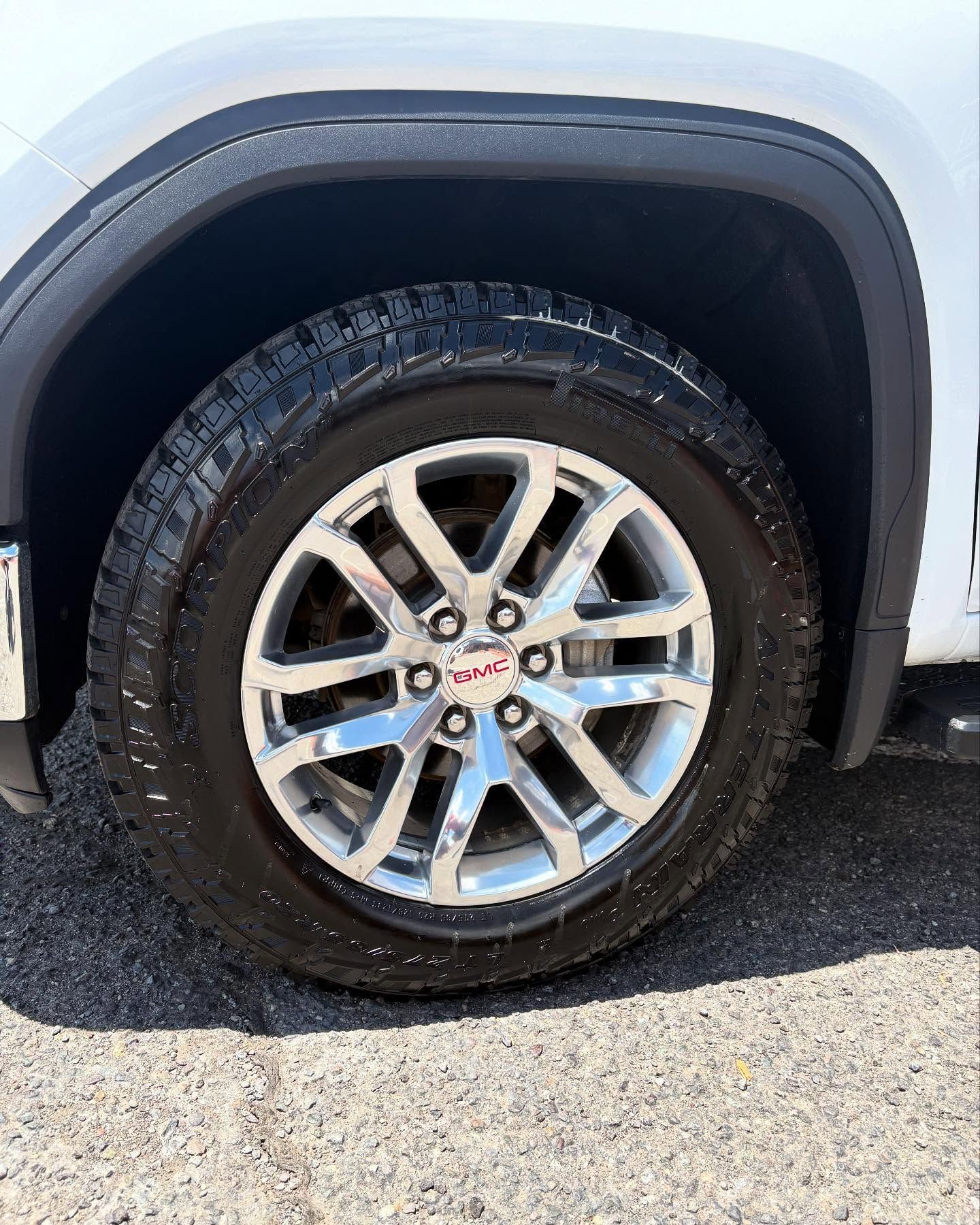 A close-up of a silver GMC multi-spoke wheel with an all-terrain tire on a white vehicle against an asphalt background.