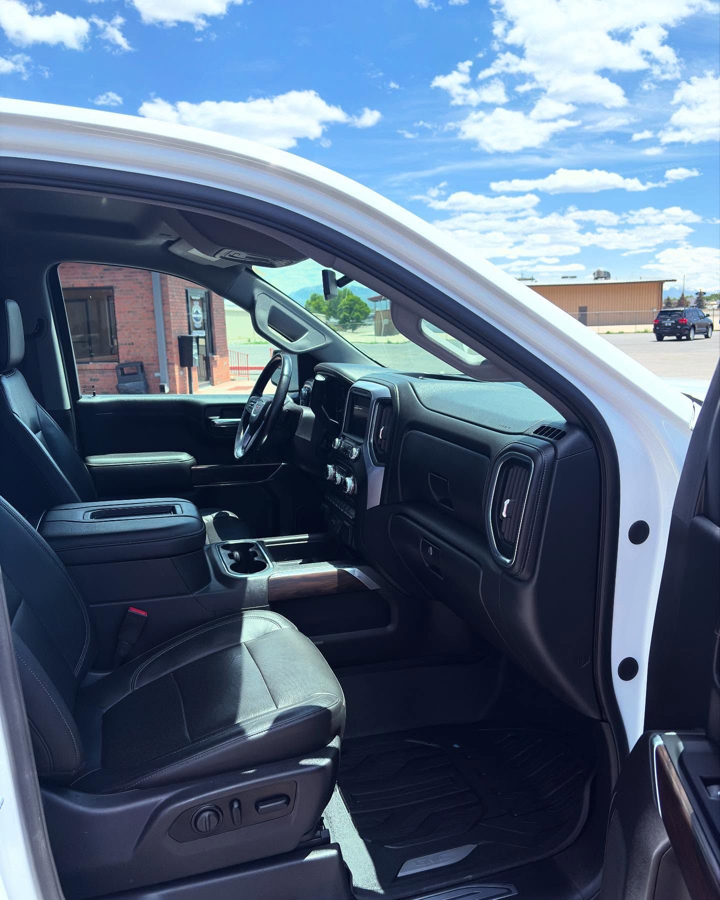 The driver's side interior of a white truck showing black leather seats, a dark dashboard, and a view of a sunny parking lot.