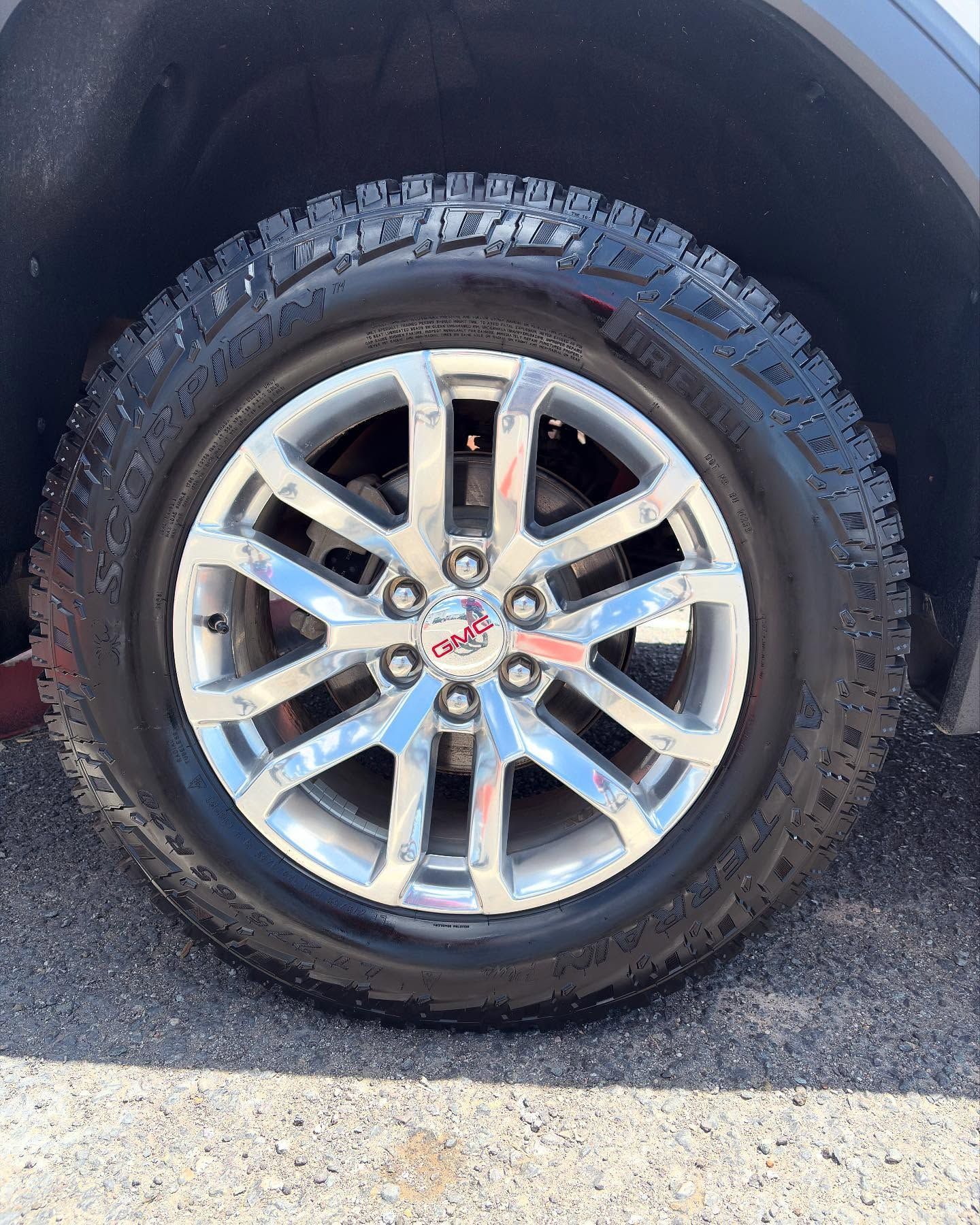 A close-up of a GMC vehicle’s chrome alloy wheel with a dark, treaded all-terrain tire, set against dark pavement.