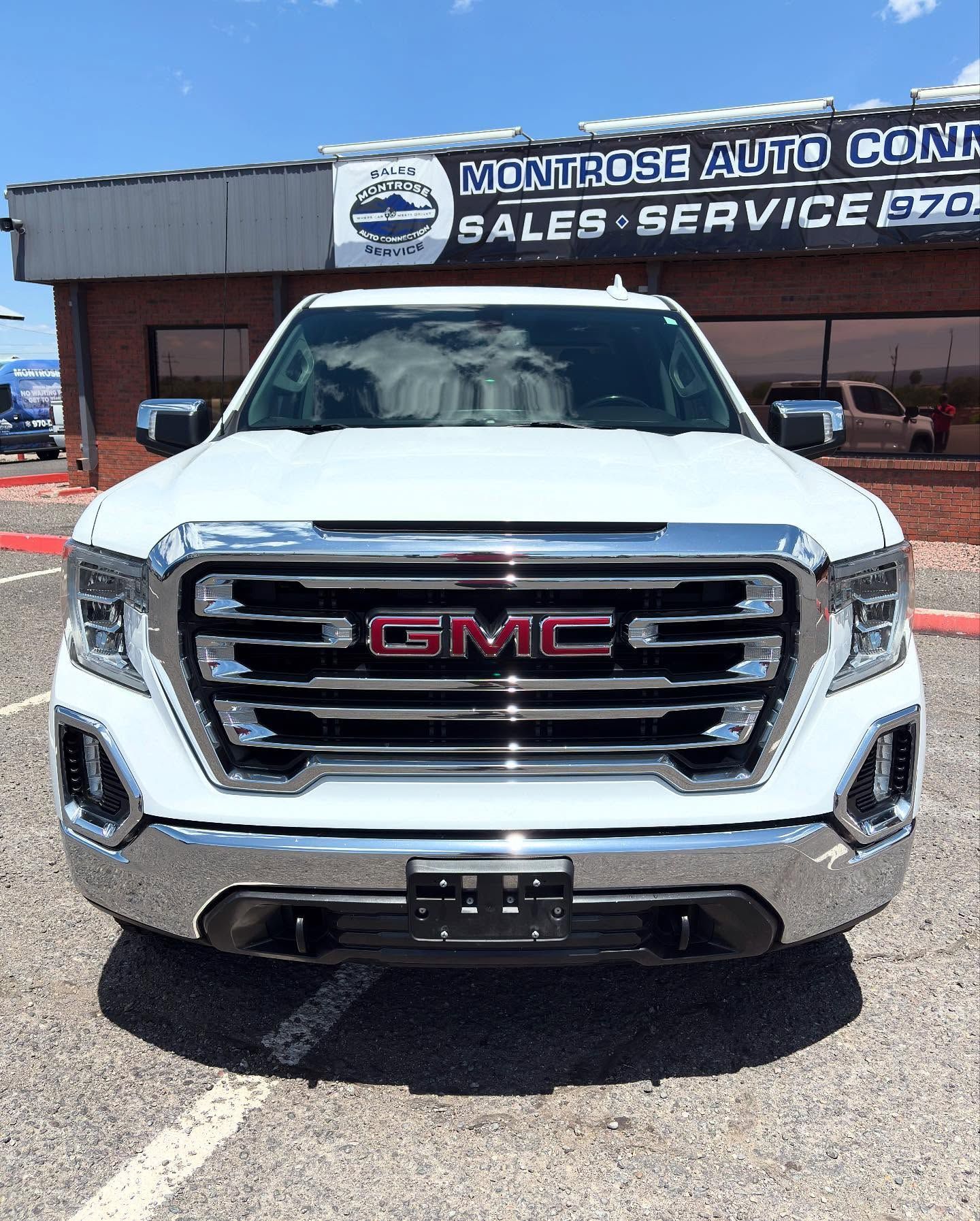 A white GMC Sierra pickup truck parked in front of a Montrose Auto sales and service building under a bright blue sky.