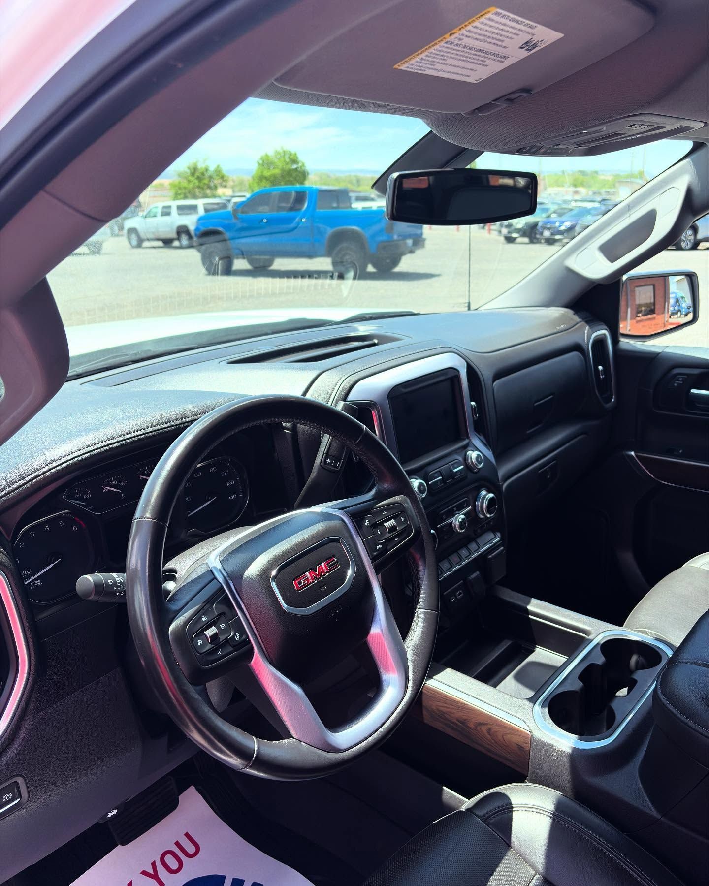 A view from inside a GMC truck cabin showing the black leather interior, steering wheel, and a sunny outdoor parking lot.