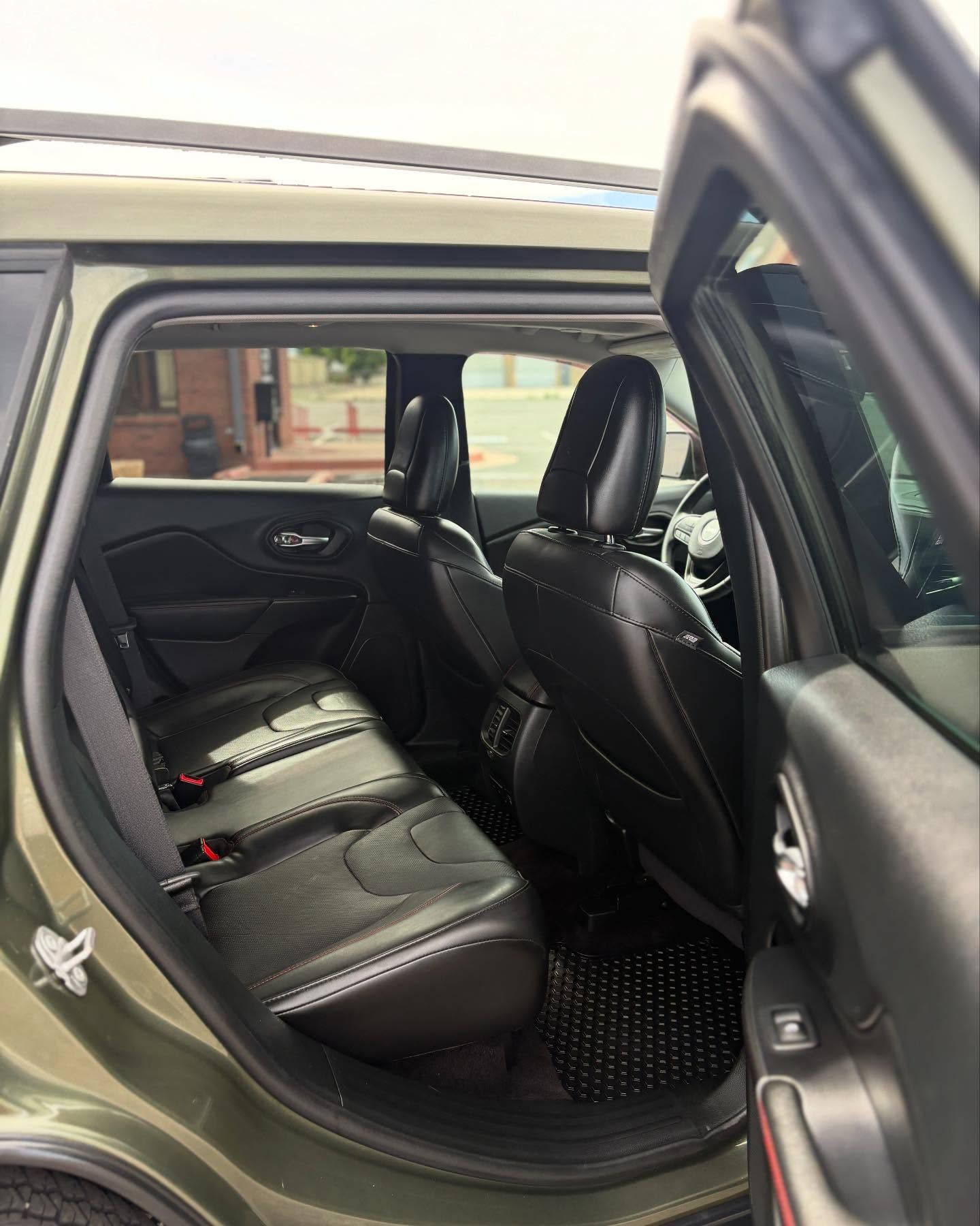 The interior of an olive green car, showing the back passenger seat with black leather upholstery and floor mats.