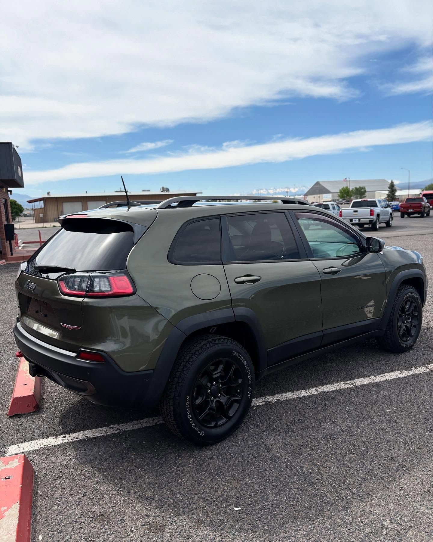 An olive green Jeep Cherokee Trailhawk parked in a gravel lot on a bright, sunny day.