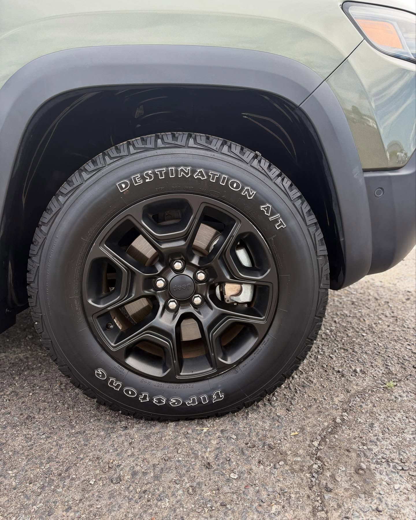A close-up of a matte black vehicle wheel with a black Firestone Destination A/T tire against a green fender.