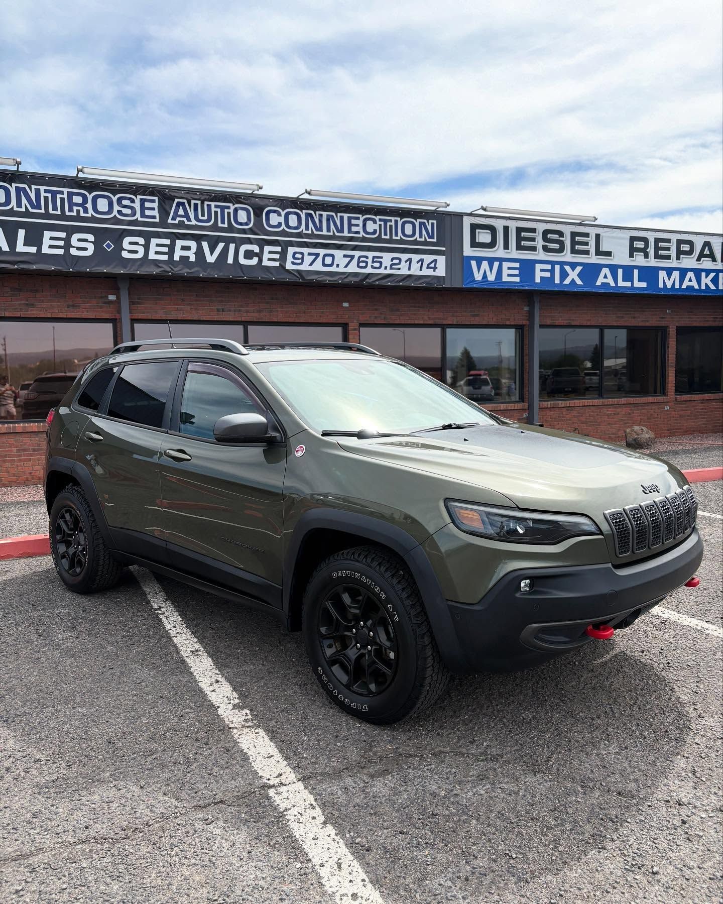 An olive green Jeep Cherokee Trailhawk parked in front of a brick building labeled Montrose Auto Connection.