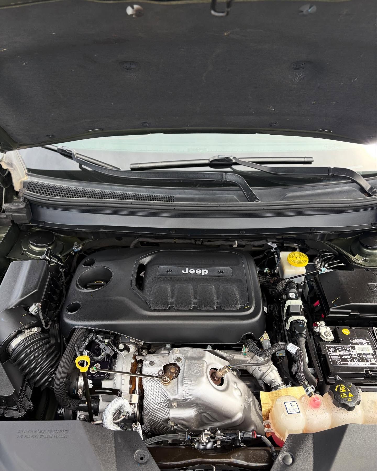The open hood of a Jeep vehicle reveals a black engine cover, a silver heat shield, and various automotive components.