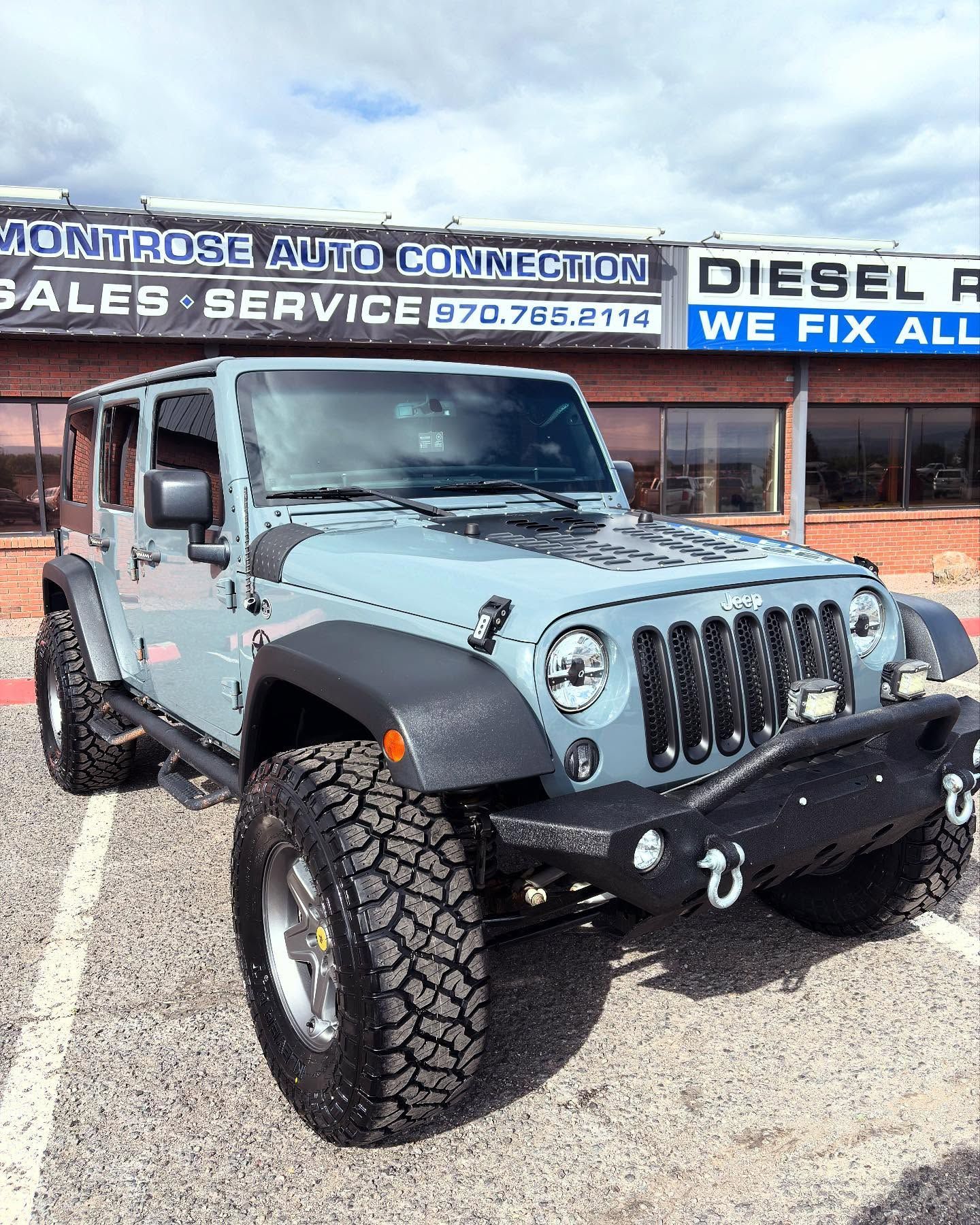 A light blue Jeep Wrangler parked in a lot in front of a Montrose Auto Connection dealership building.
