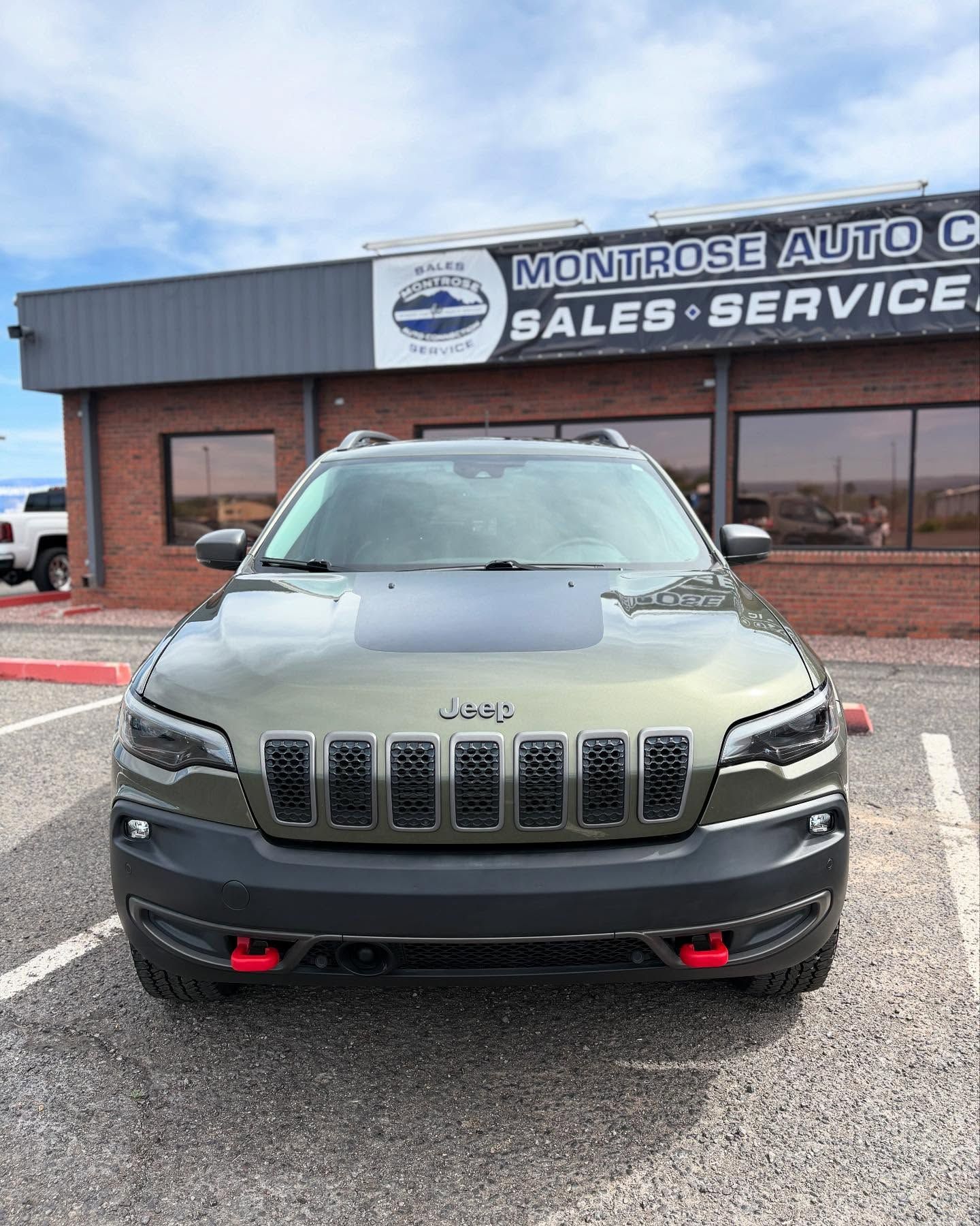 An olive-green Jeep Cherokee Trailhawk parked in front of a brick auto dealership building under a cloudy sky.