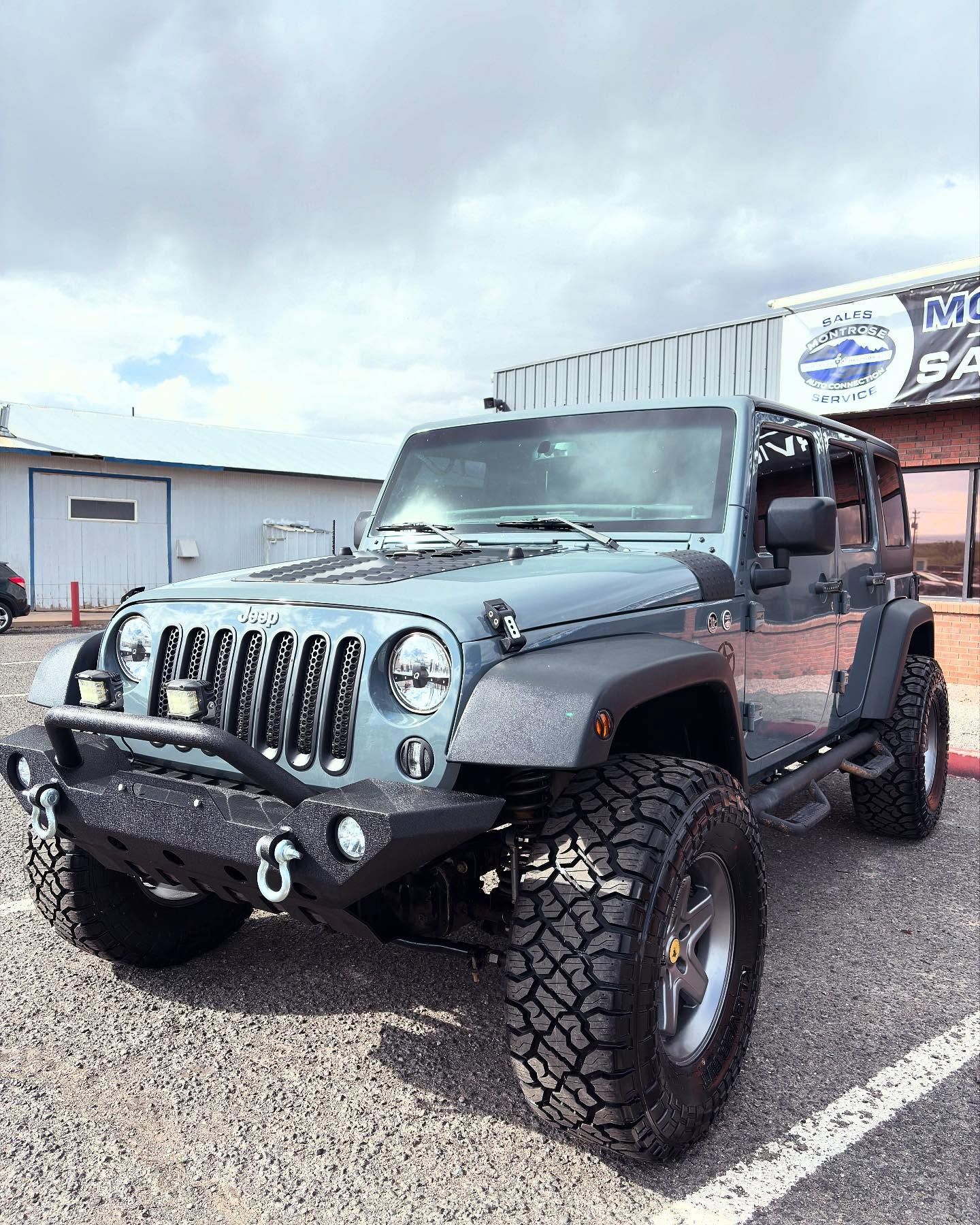 A grey Jeep Wrangler modified with an off-road bumper, winch, and large tires, parked on a gravel lot under a cloudy sky.