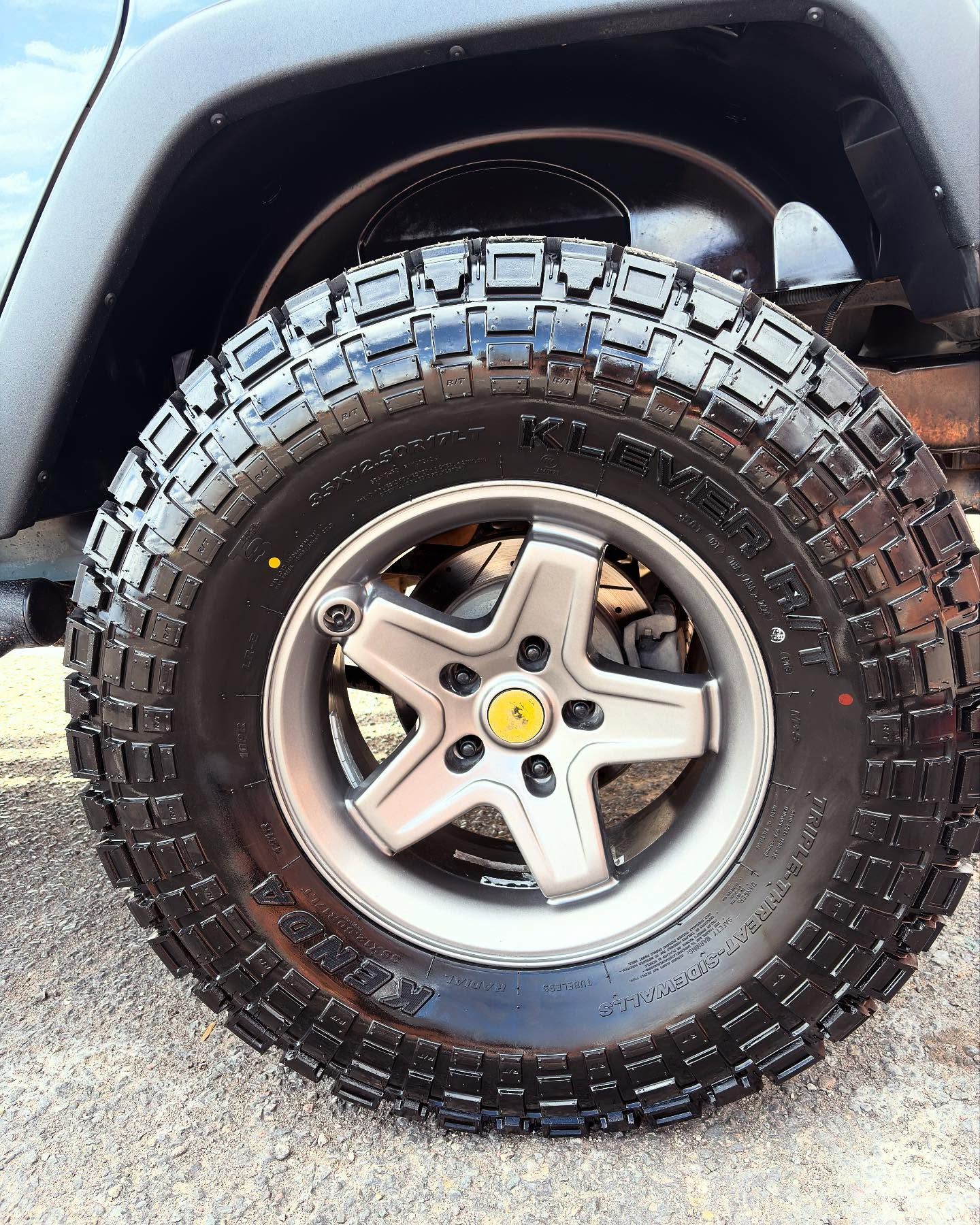 A close-up of a dark, rugged off-road tire on a silver five-spoke alloy wheel with a yellow center cap.
