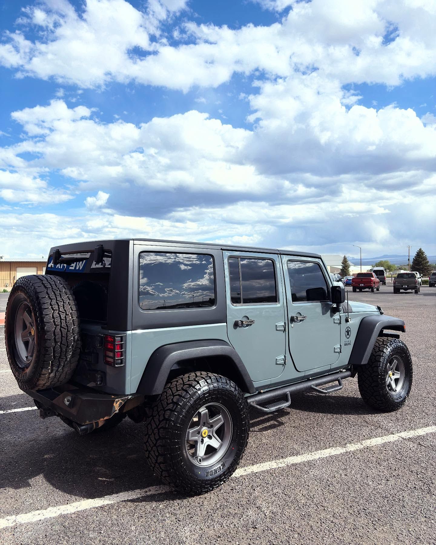 A gray Jeep Wrangler parked on a gravel lot under a bright blue sky with large, puffy white clouds.
