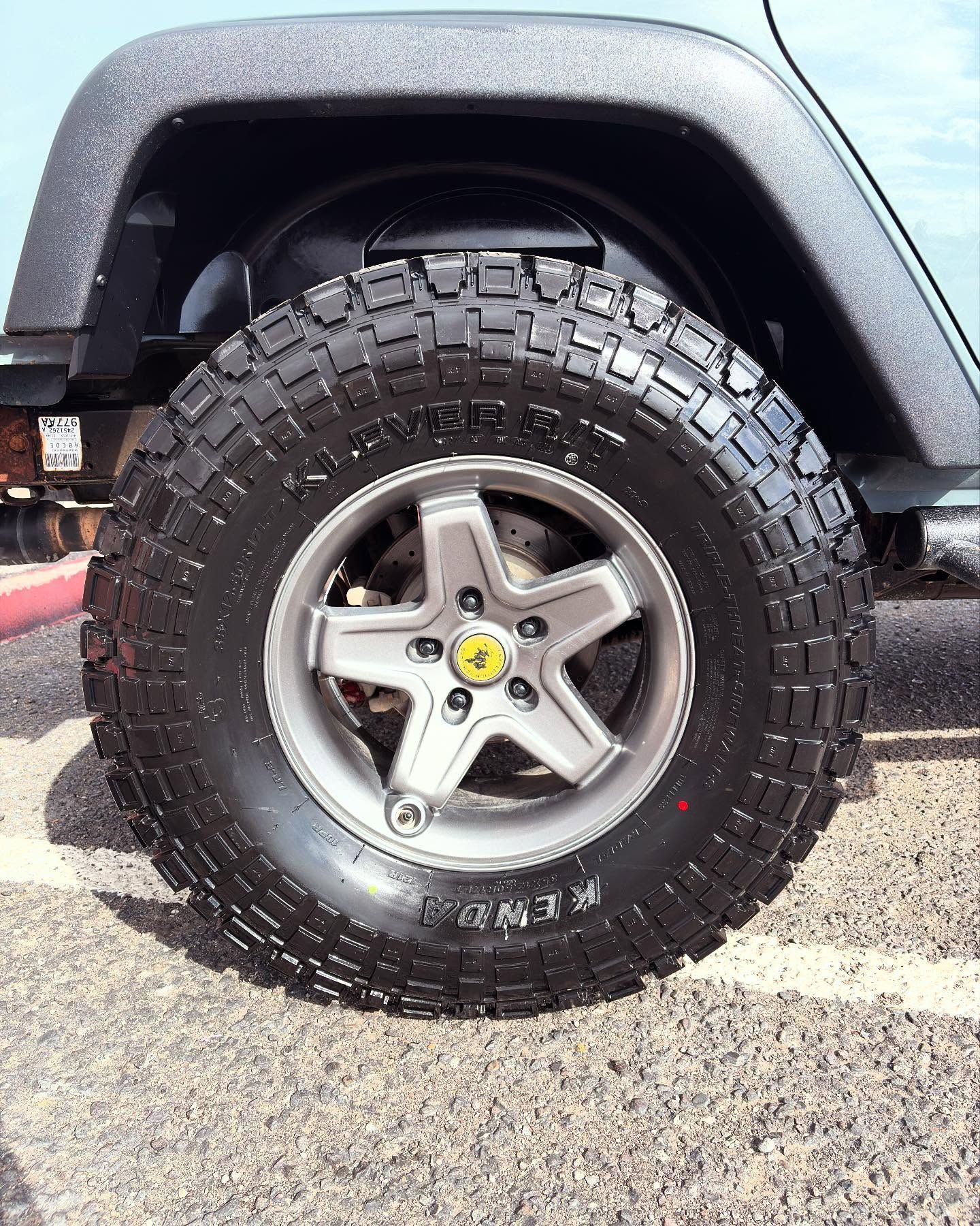 A close-up of a rugged, off-road vehicle tire with a textured tread pattern on a five-spoke gray wheel in a parking lot.