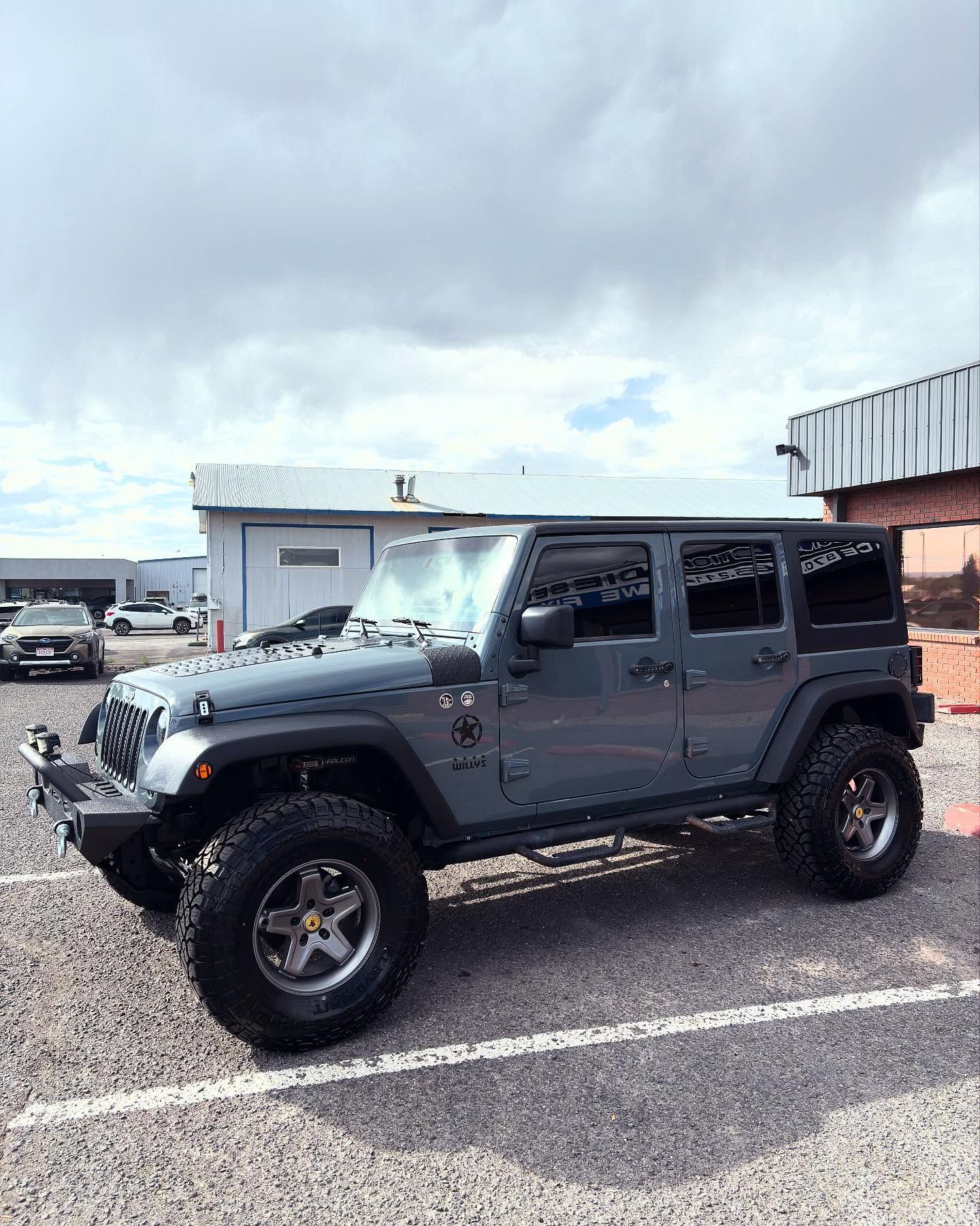 A dark gray four-door Jeep Wrangler with large off-road tires parked in a gravel parking lot under a cloudy sky.