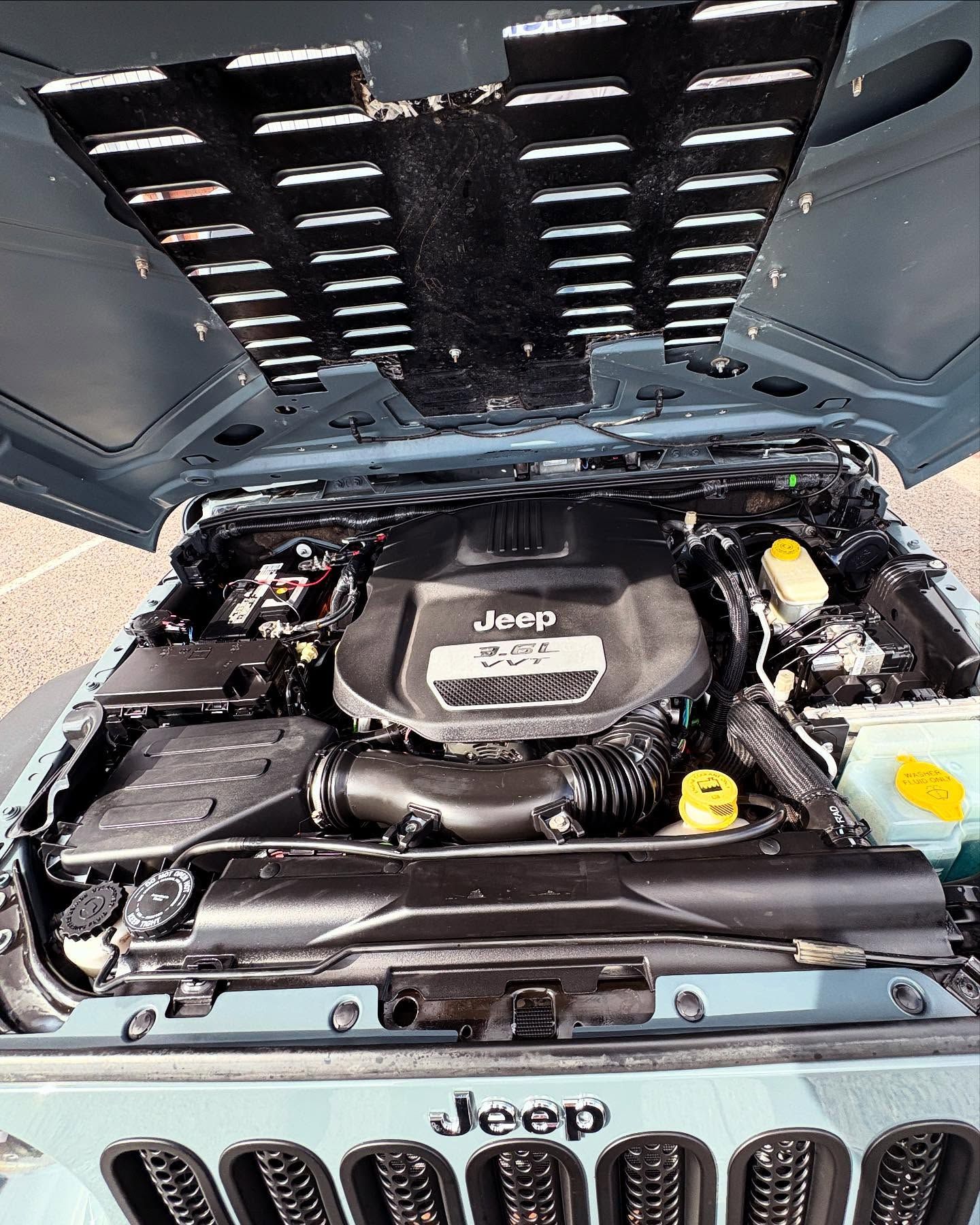 The open hood of a gray Jeep reveals its engine bay, including the engine cover with the Jeep logo and a vented hood.