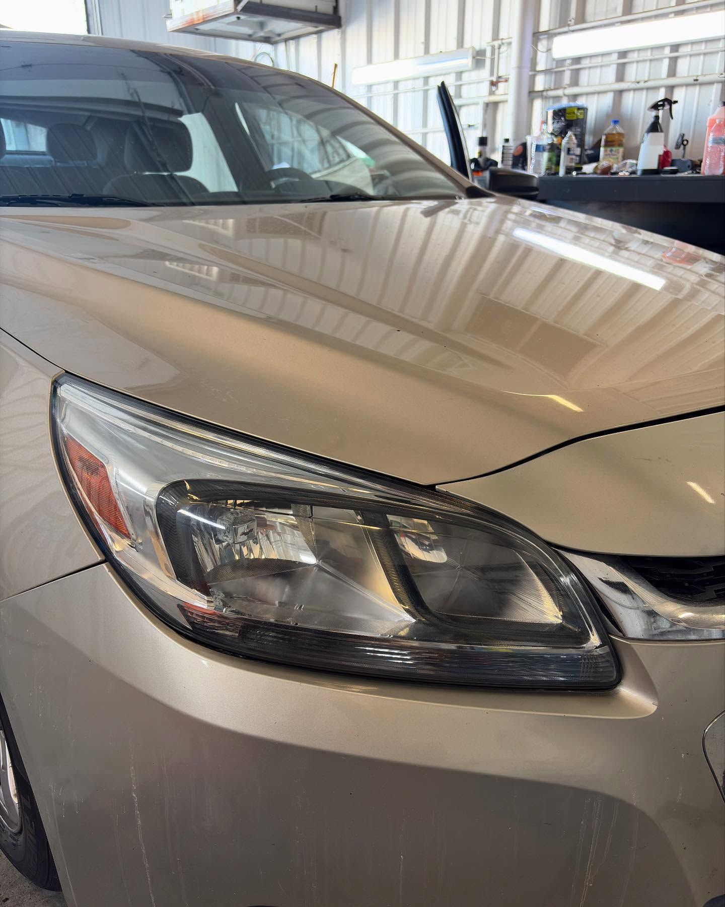 A close-up view of the front passenger side headlight and hood of a beige car parked in an auto repair shop.