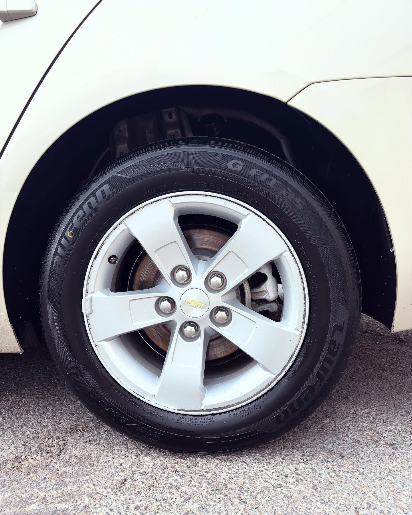 A close-up of a silver, five-spoke alloy wheel with a Chevrolet emblem, mounted with a black tire on a light-colored car.