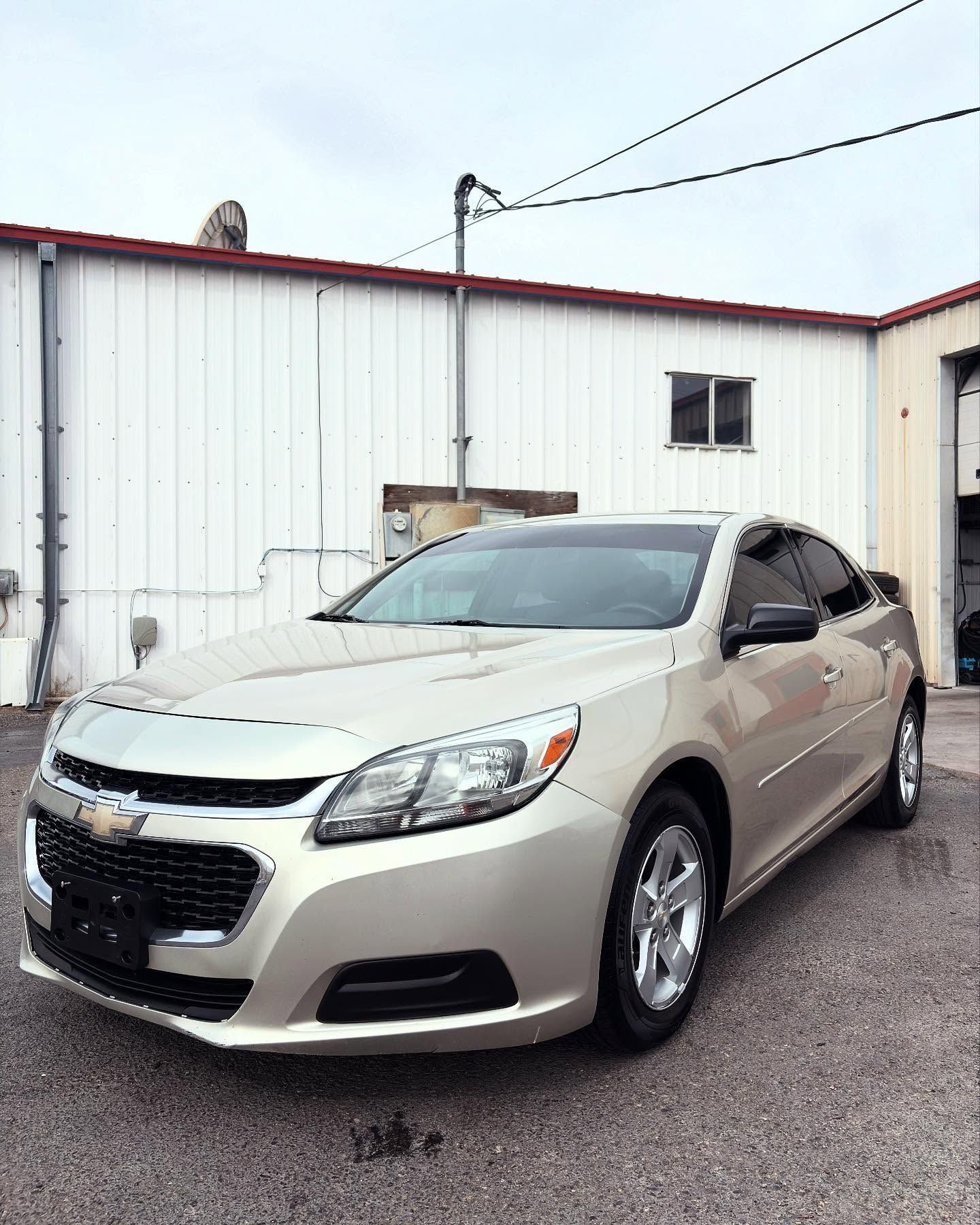 A light gold Chevrolet Malibu sedan parked on a gravel lot in front of a white metal industrial building.