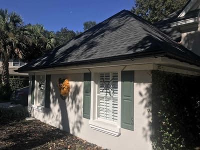 White cottage with dark roof, green shutters, and a front window, surrounded by trees in sunlight