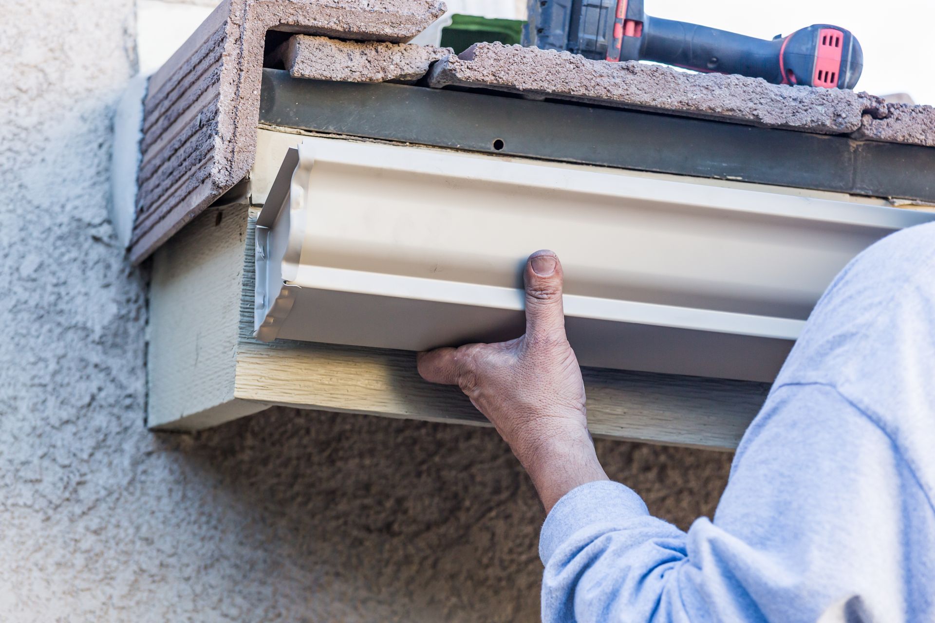 Person installing a white window blind under an outdoor ledge with a drill nearby