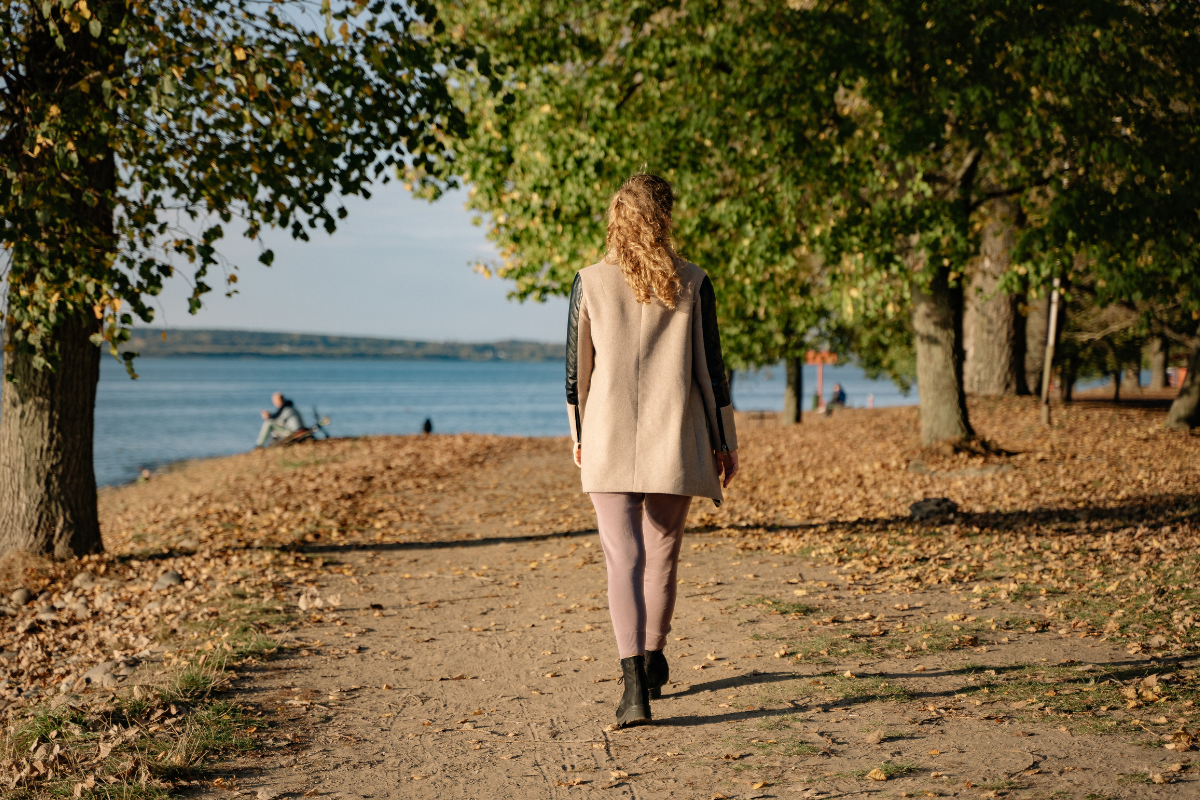 A woman walks alone along a tree-lined park path, viewed from behind.