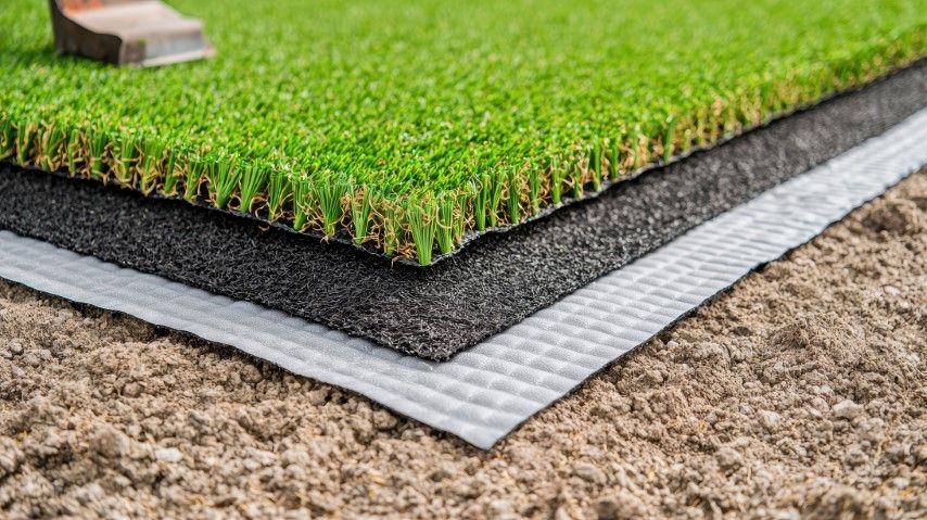 Layers of artificial turf: green grass, black padding, white base, on brown ground.