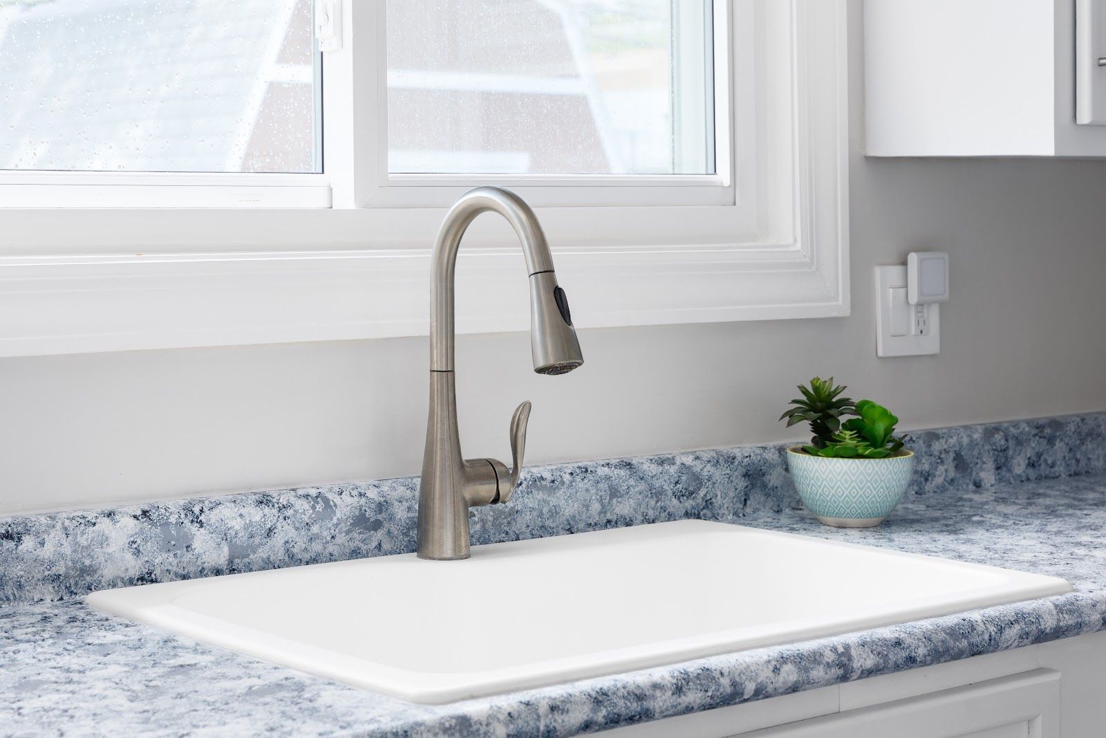 Kitchen sink with faucet, white basin, blue speckled countertop, small plant, window.