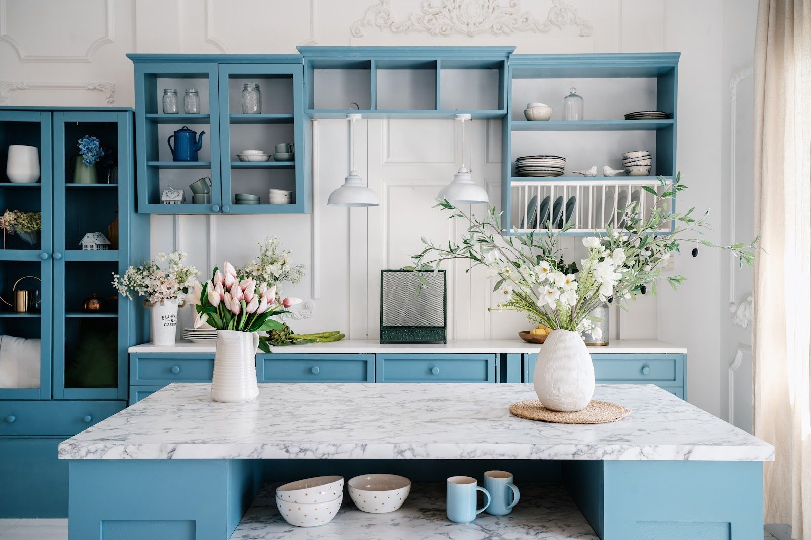 Blue kitchen with island, marble countertop, open shelving, white walls, and floral arrangements.