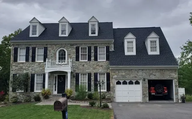 A man is standing in front of a large stone house with a black roof.