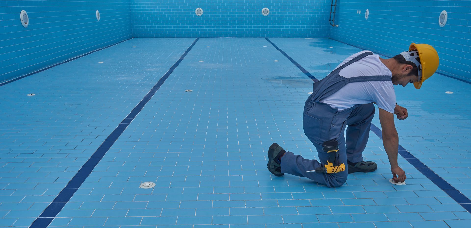 Technician kneels to inspect pool drain in empty tiled swimming pool.