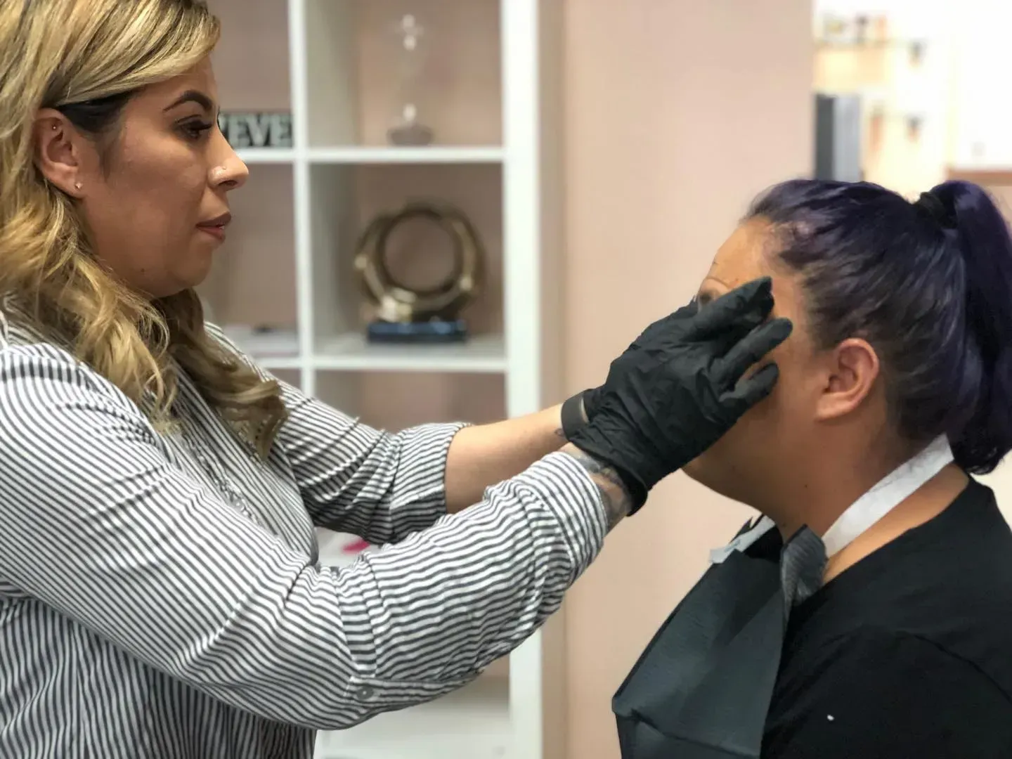 A woman with purple hair is getting her eyebrows done by a woman wearing black gloves.