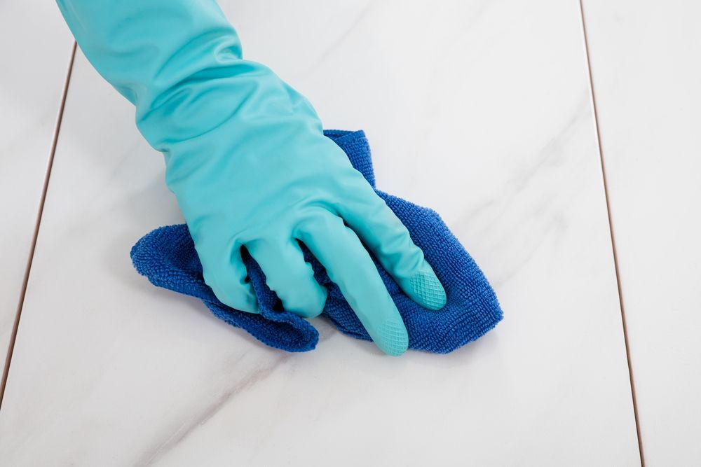 A gloved hand wiping a white tile floor with a blue microfiber cloth.