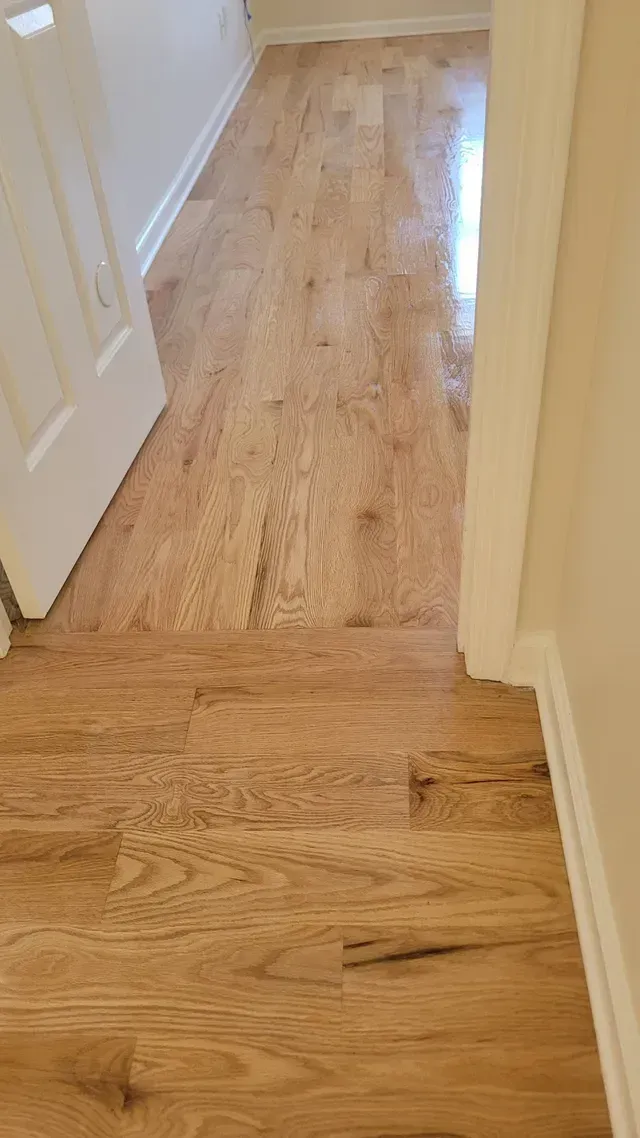 Light wood flooring in a hallway with a white door on the left and cream-colored walls.
