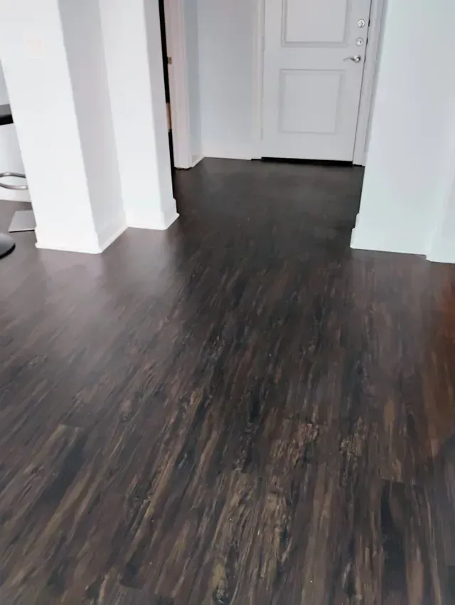 Dark wood-look flooring in a room with white walls and door; a doorway leads to another area.