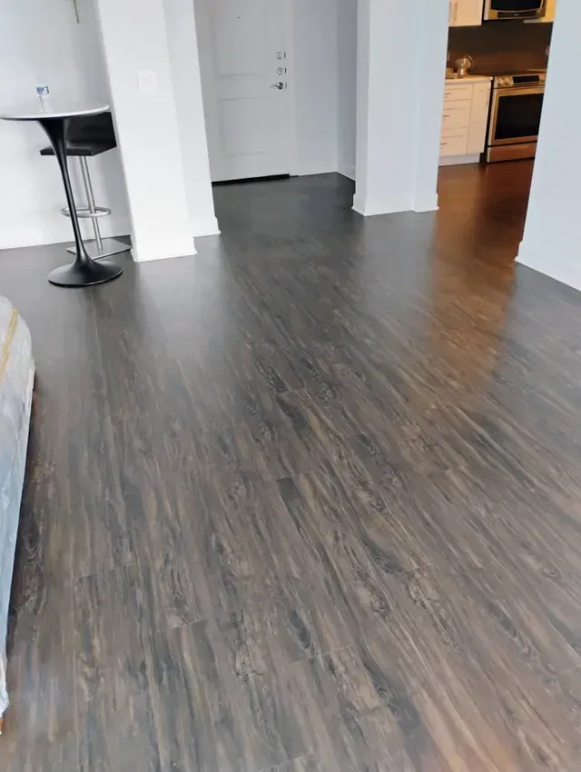 Dark wood-look flooring in an apartment, leading towards a white door and kitchen