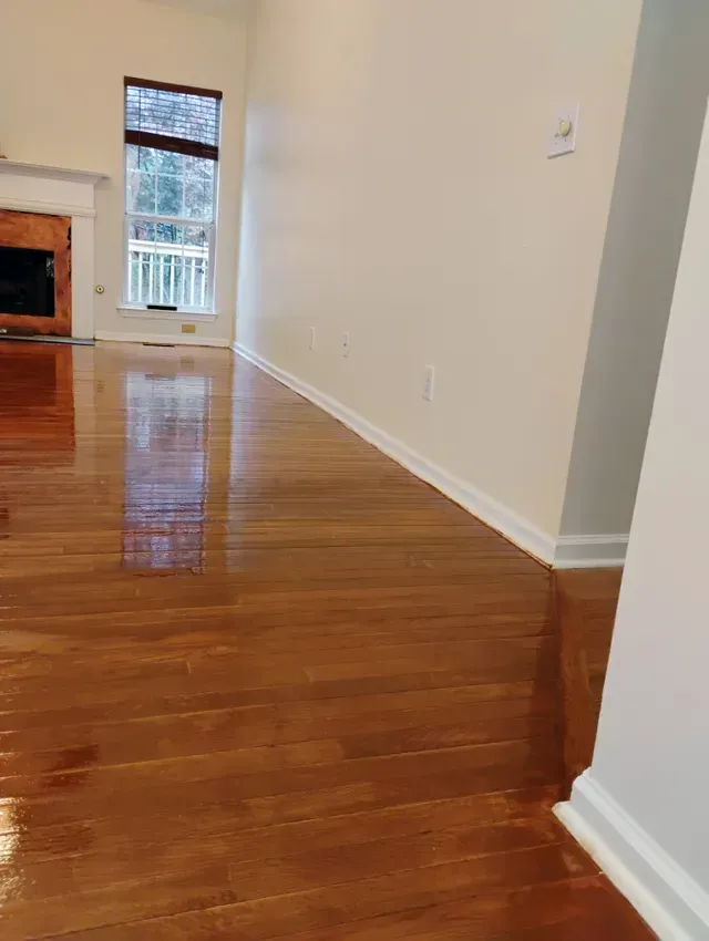 Shiny wood floor in a room with a tall window, fireplace, and off-white walls.
