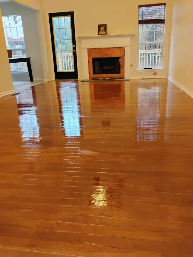 Shiny, newly-finished wooden floor reflecting a living room's windows, fireplace, and door.