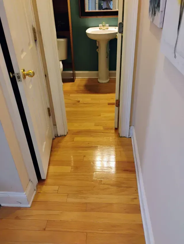 Narrow hallway with wooden floor, leading to a bathroom with a pedestal sink and dark green wall.