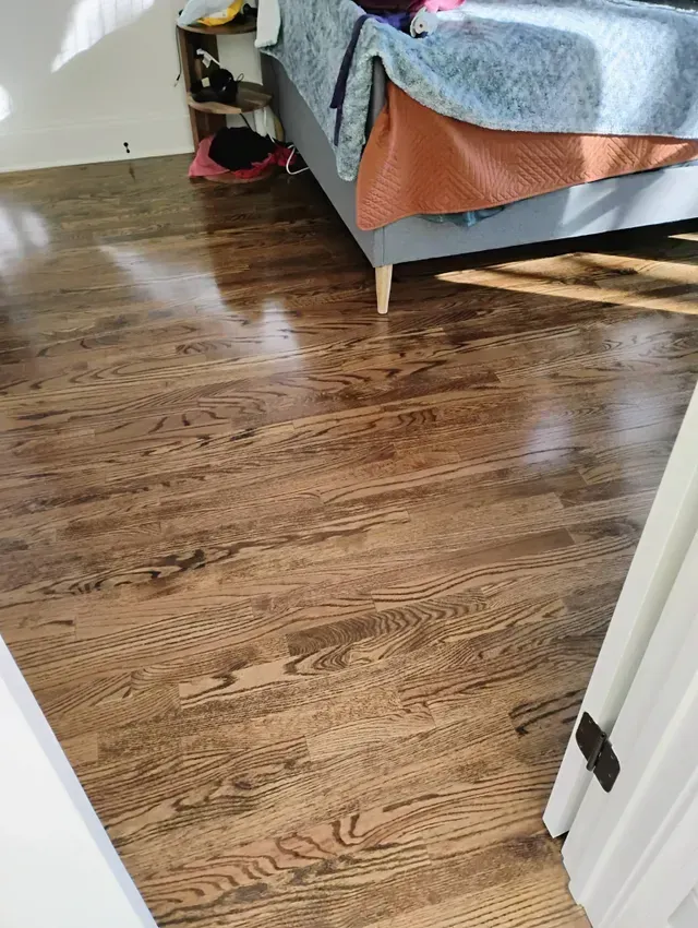 Brown hardwood floor in a room, with a bed and objects visible.