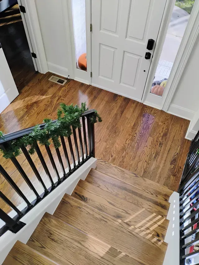 Wooden staircase leads to a hallway with hardwood floors, front door visible. Black railing with garland.