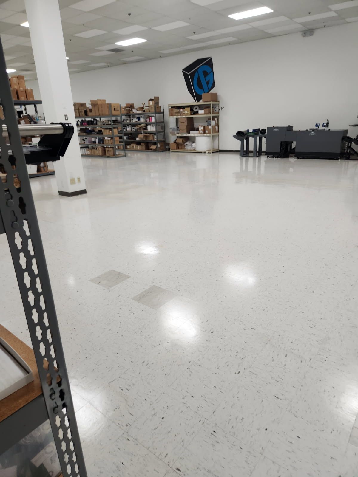 Large, white-floored warehouse; shelving with boxes, printing equipment, logo on wall, tall white pillar.