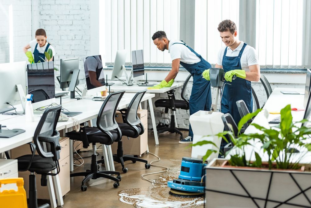 Office cleaning crew wiping desks and scrubbing floor with equipment.