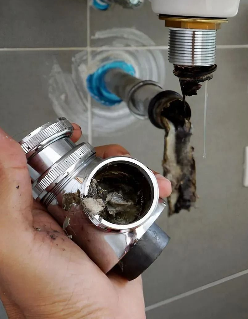 Hand holding a clogged chrome drain fitting beneath a bathroom sink with exposed pipes