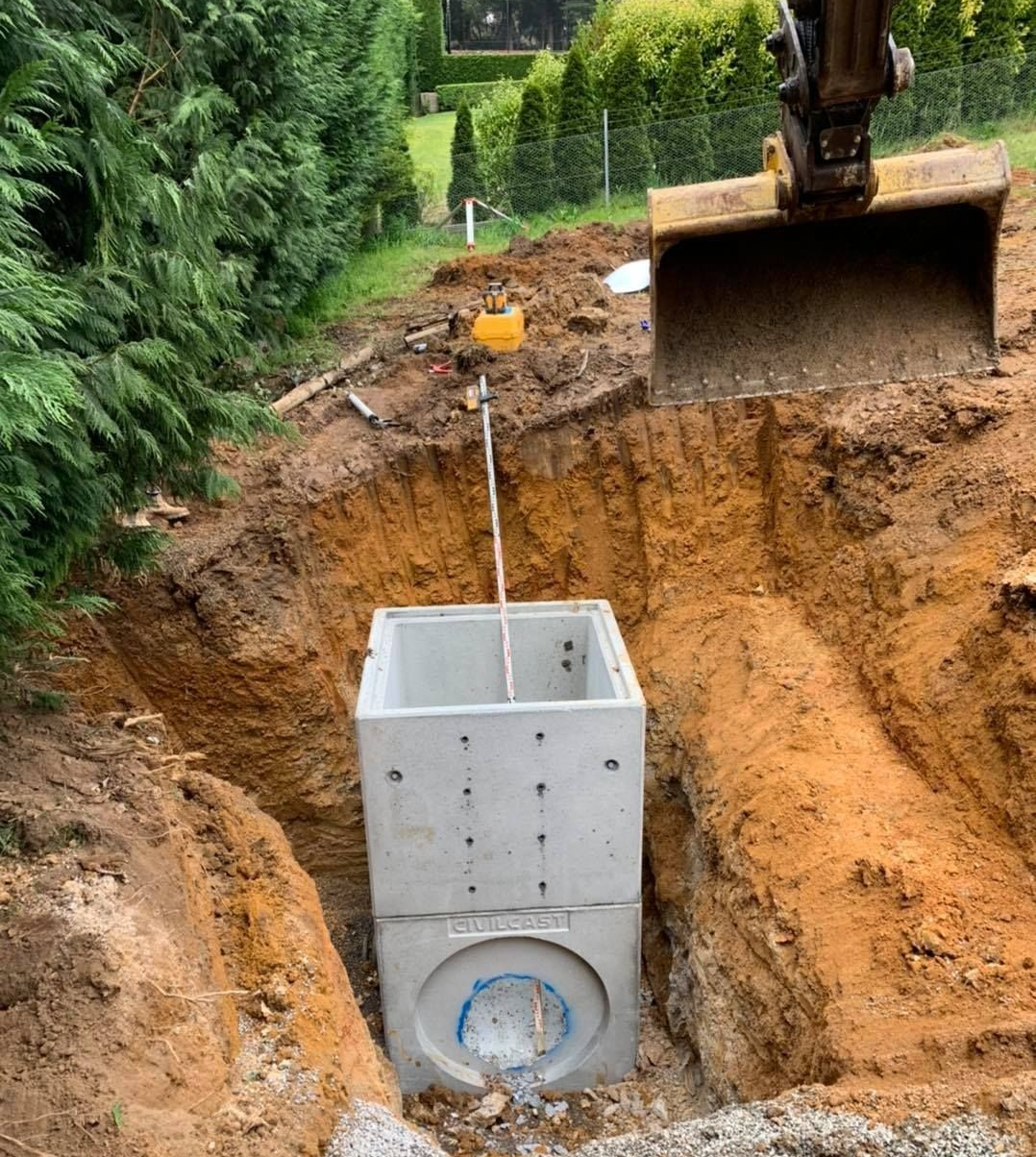 Excavator placing a concrete utility chamber into a dug trench beside a hedge-lined yard