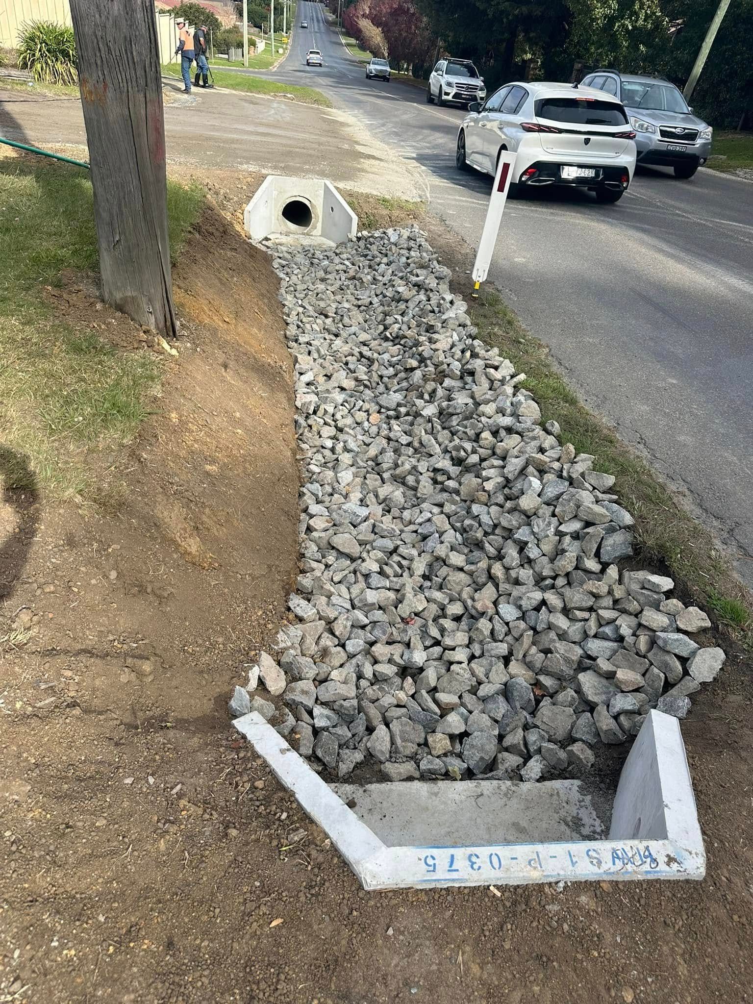 Rock-filled roadside drainage trench beside a road, lined with white markers and a culvert pipe end.