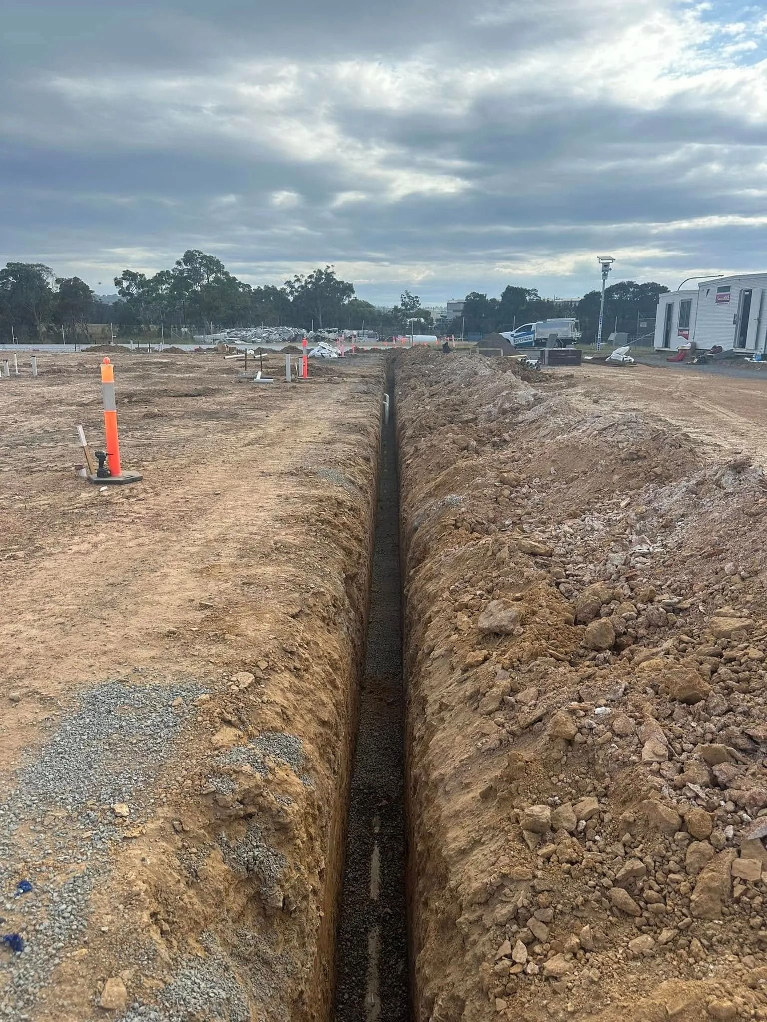 Long trench cut through dirt at a construction site under cloudy skies, with orange cones and equipment nearby